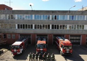 Septiņi ugunsdzēsēji glābēji stāv pie trīs dažādām ugunsdzēsēju glābšanas autotehnikām, ietur klusuma brīdi un rokās tur savas ķiveres