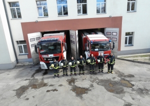 Septiņi ugunsdzēsēji glābēji stāv vienā rindā. Rokās tur savas ķiveres. Aiz viņiem redzamas divas ugunsdzēsēju glābēju automašīnas