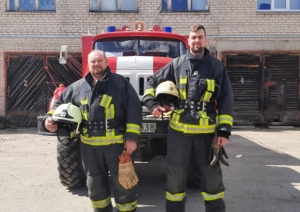 Ģērbušies savos aizsargtērpos un turot rokās ķiveri stāv divi ugunsdzēsēji glābēji. Viņiem fonā redzama ugunsdzēsības un glābšanas dienesta automašīna