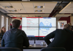 Sanāksmju zāle. Priekšplānā divi vīrieši ar mugurpusi. Skats uz ekrānu, kurā tiek rādīta projekta prezentācija.