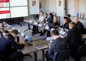 Sanāksmju zāle. Pie galda projekta pārstāvji. Pretī ekrāns ar prezentāciju.